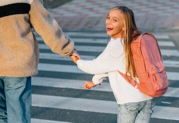 lachendes Mädchen mit Rucksack geht an der Hand eines Elternteils über einen Zebrastreifen