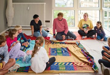 Mehrere Kinder und drei Erwachsene sitzen in einem Raum auf dem Boden und reden miteinander