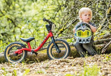 Kleines Kind sitzt auf einem Baumstamm, auf dem Schoß ein Fahrradhelm, neben ihm ein Laufrad