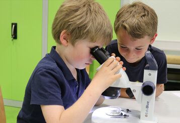 Zwei Jungs sitzen an einem Mikroskop mit einer Petrischale,  einer schaut ins Mikroskop rein, der andere guckt zu