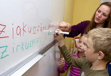 Eine Lehrerin und zwei Kinder stehen an einem White Board, ein Junge schreibt etwas auf das Board