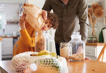 Junge kippt Nudeln aus einer Papiertüte in einen Glasbehälter, Vater steht daneben, im Hintergrund Küche