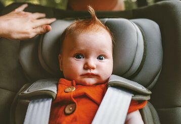 Ein Baby sitzt im Auto in einem Kindersitz, guckt lustig und hat eine Haartolle