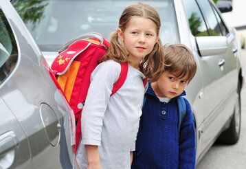 Zwei Schulkinder stehen zwischen parkenden Autos und versuchen, die Straße zu überqueren