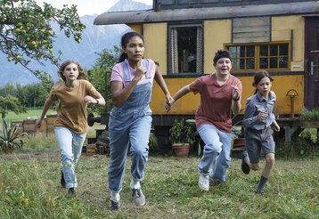 Vier Kinder rennen auf einer Wiese, im Hintergrund ein Wohnwagen und Berge